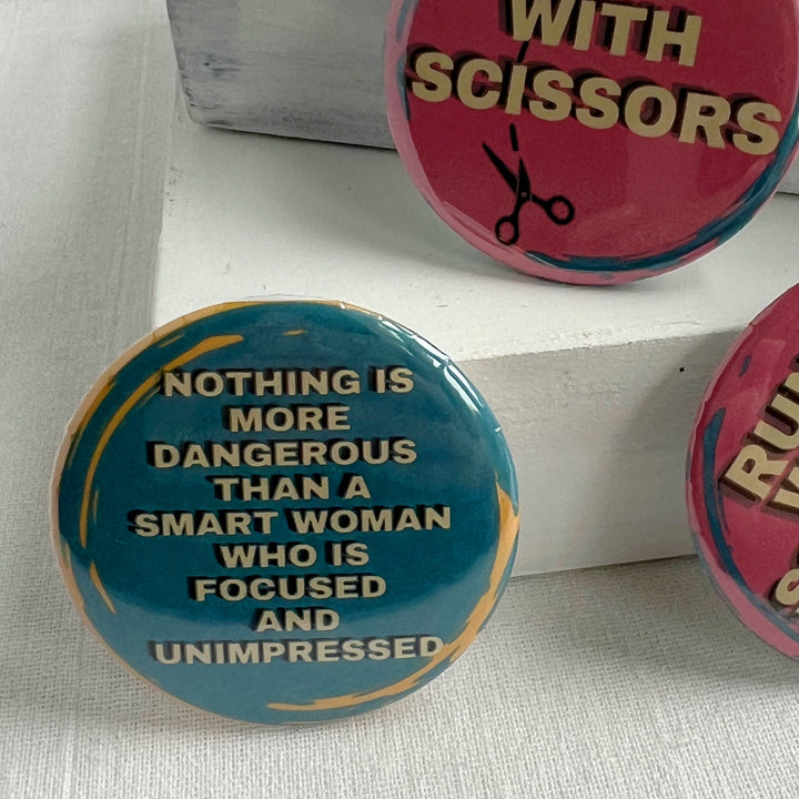 round pinback button with a blue backgroun, yellow streaks and the words, "Nothing is more dangerous than a smart woman who is focused and unimpressed."