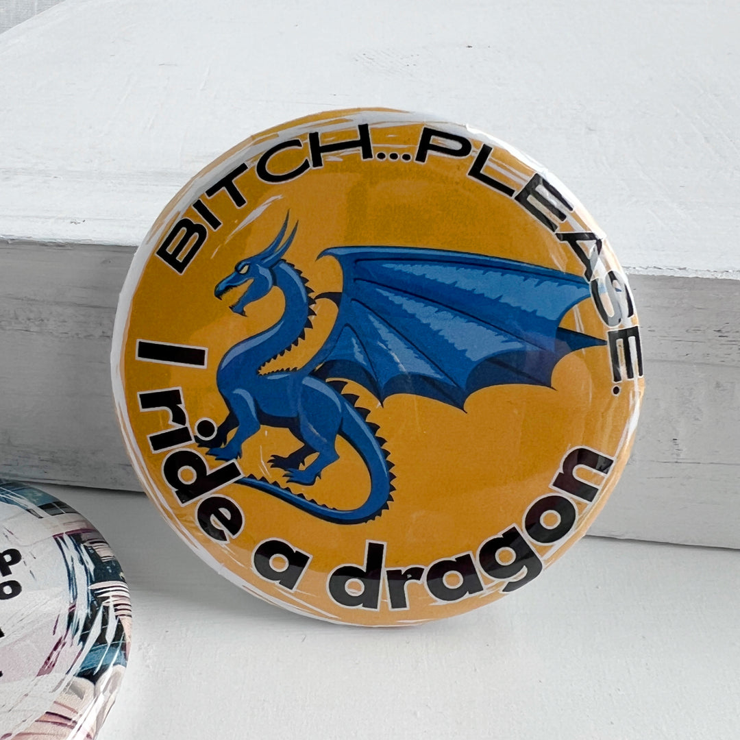 round pinback button with a yellow background, a blue dragon and the words, "bitch..please. I ride a dragon" in black lettering.