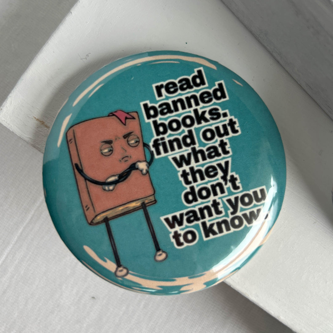 round pinback button with a teal blue background, a cartoon figure of a book with an angry face, and the words, "read banned book. find out what they don't want you to know."