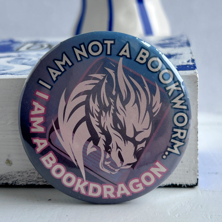 round pinback button with an ombre colored background with blues/pinks/purples, with the words, "I am not a bookworm..I am a book dragon" surrounding a light pink dragon figure on top of a book.