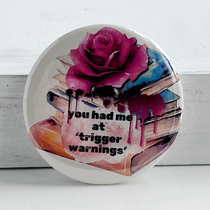 Round pinback button with a white background, a stack of colorful books with a rose on top and some blood dripping with the words, "You had me at trigger warnings."