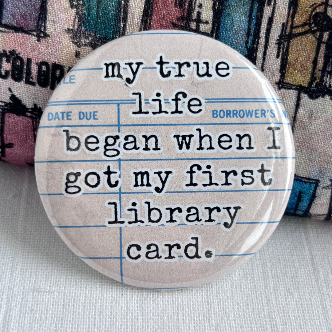 round pinback button with a library checkout card background and the words, "my true life began when I got my first library card."