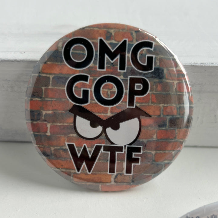 A round pin back button with a red brick background, with the letters, OMG GOP WTF and an angry face.