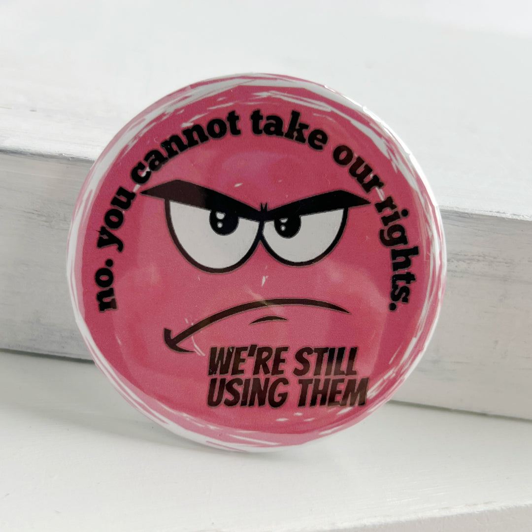 round pinback button with a pink background and the words, "no. you cannot take our rights. We're still using them."  The words surround an angry face.