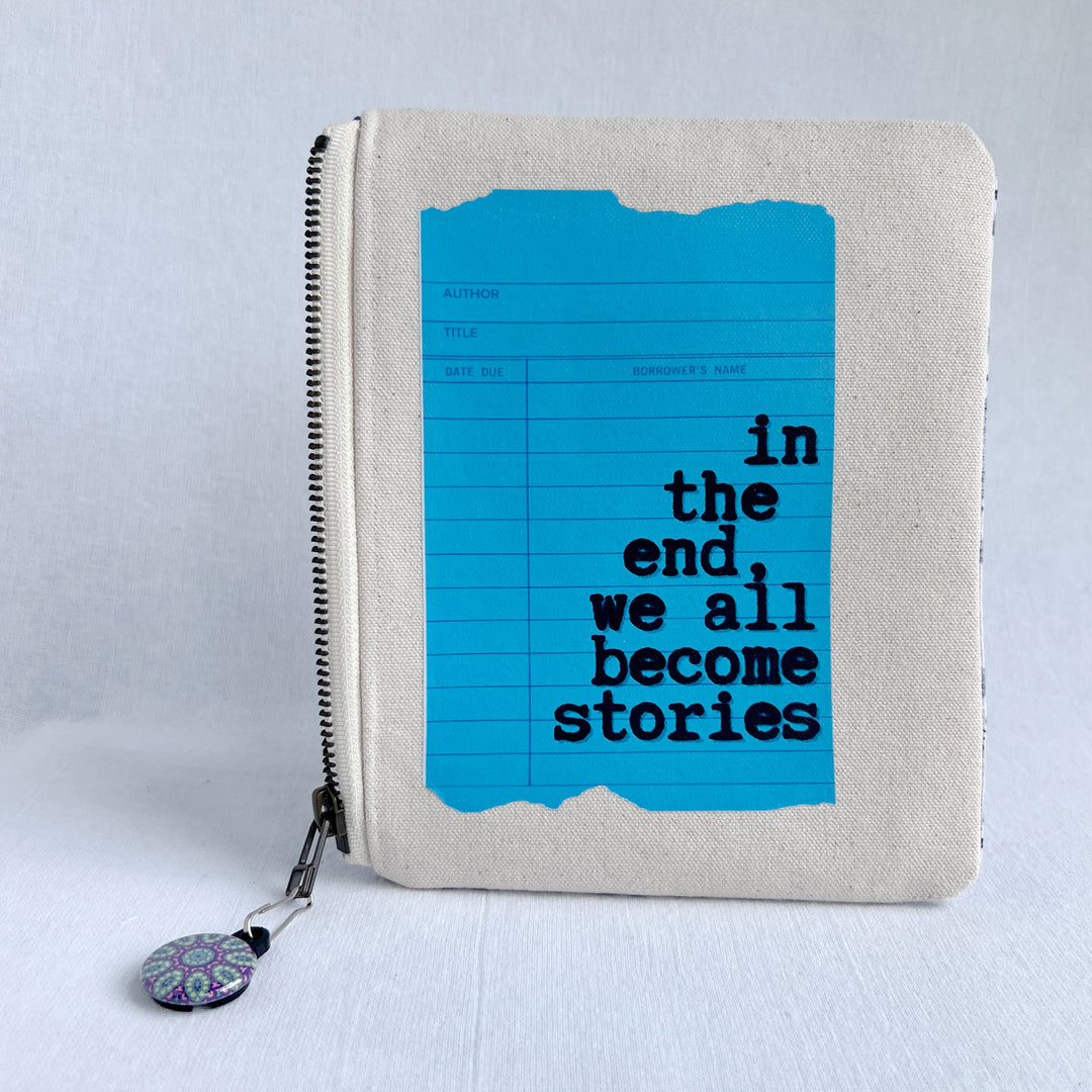 padded canvas zip bag with a blue library card and the words, in the end we all become stories plus a colorful zipper pull and lining