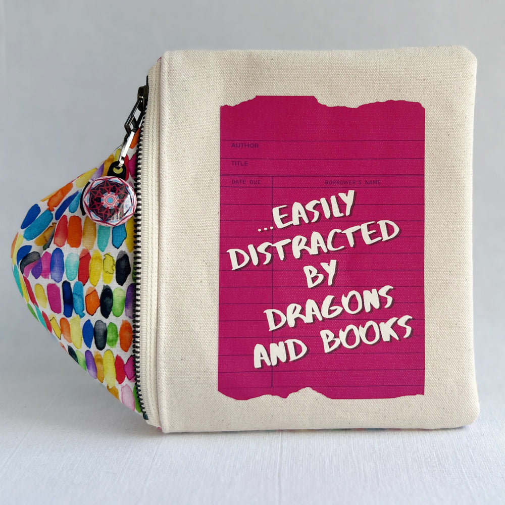 padded canvas zip bag with a coral red library card and the words, easily distracted by books and dragons, plus a colorful zipper pull and lining