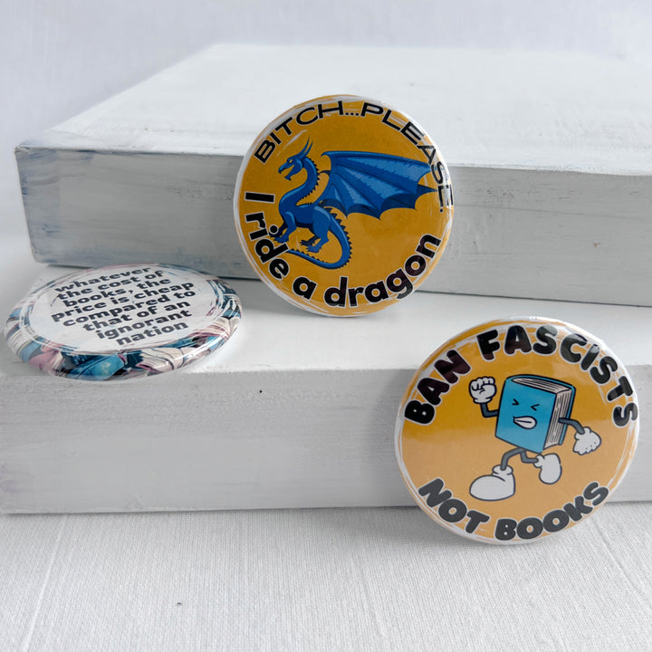 round pinback button with a yellow background, a blue dragon and the words, "bitch..please. I ride a dragon" in black lettering. Additional round pinback with a cartoon of an angry book and the words, "ban fascists, not books."