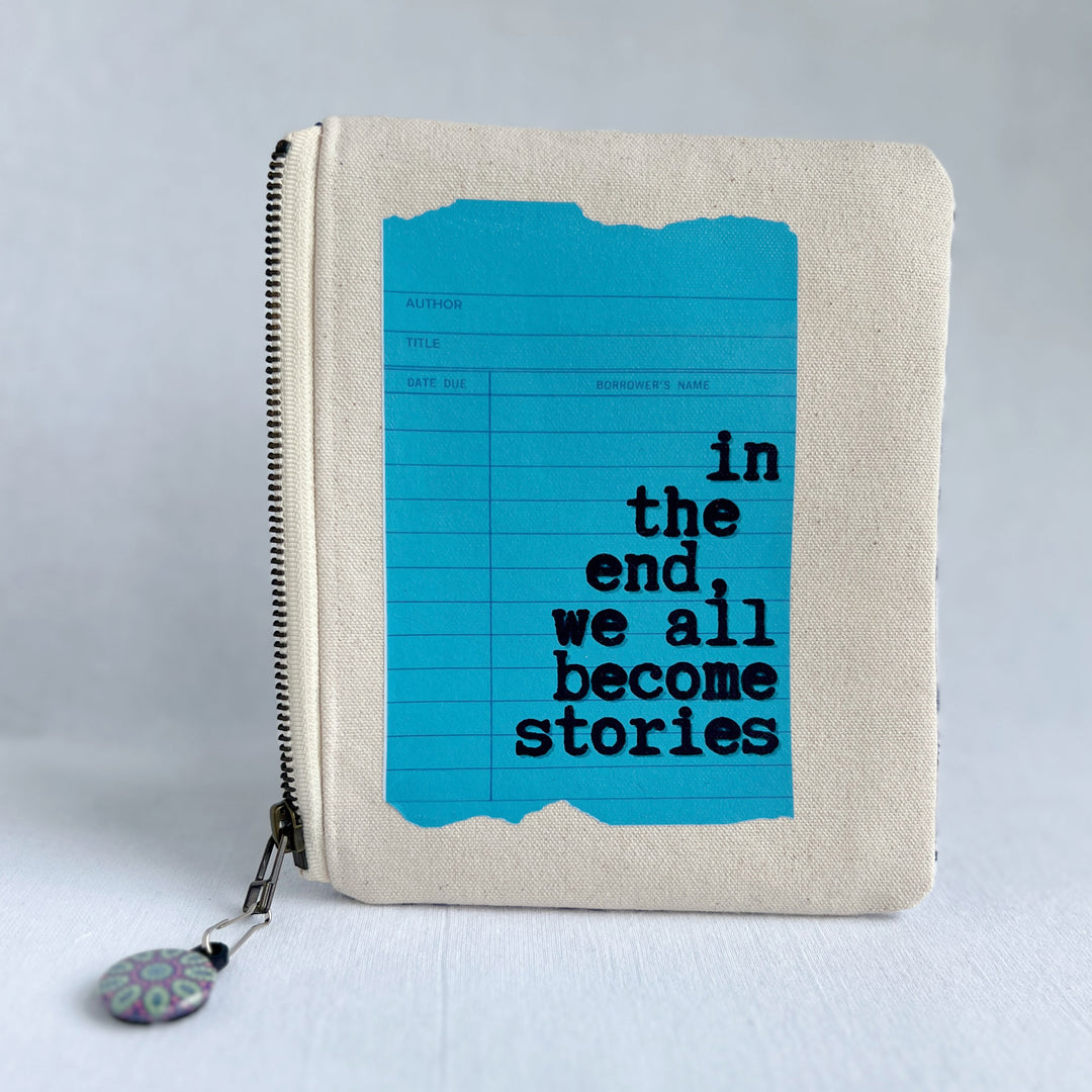 padded canvas zip bag with a blue library card and the words, in the end we all become stories plus a colorful zipper pull and lining