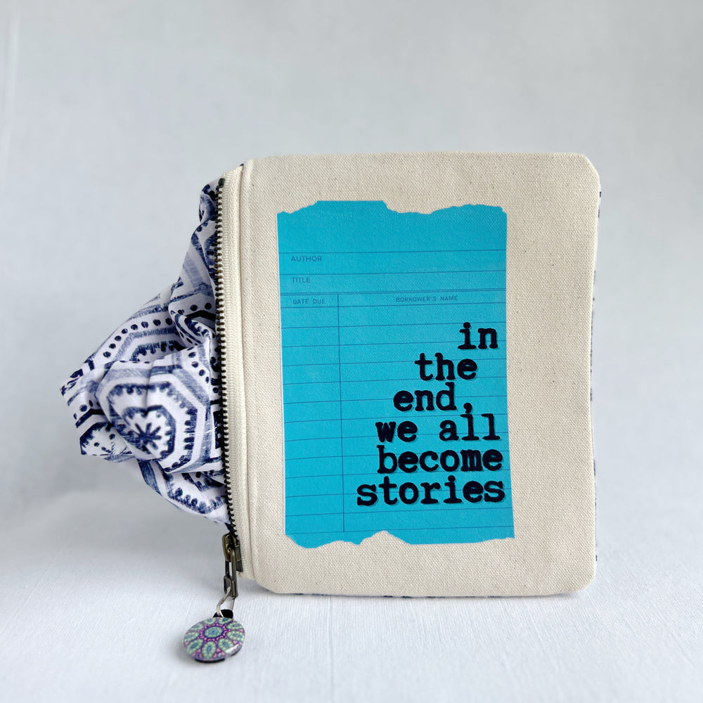 padded canvas zip bag with a blue library card and the words, in the end we all become stories plus a colorful zipper pull and lining