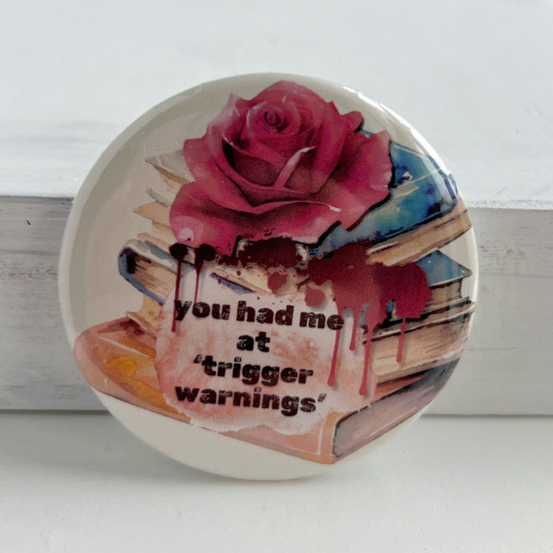 Round pinback button with a white background, a stack of colorful books with a rose on top and some blood dripping with the words, "You had me at trigger warnings."