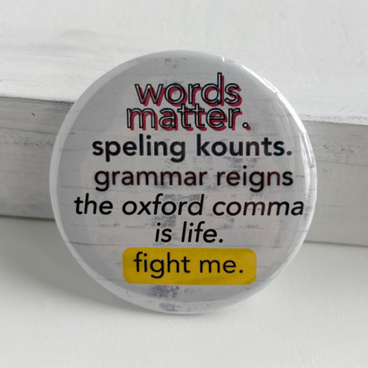 a round pin back button with a white background and the words, "words matter. speling kounts. grammar reins. the oxford comma is life. fight me."