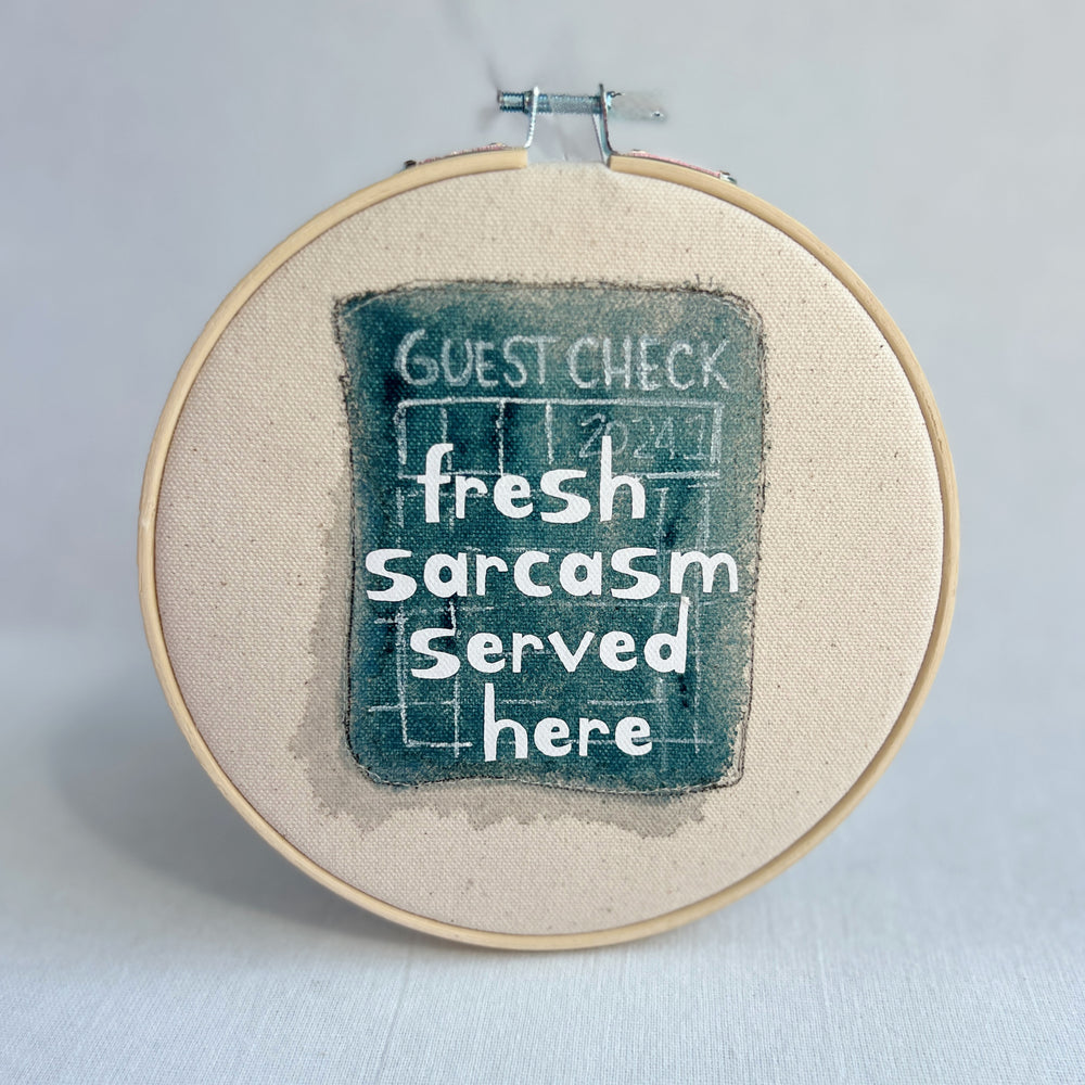 original hoop art piece with a faded guest check ticket painted in mayan blue, with the words, fresh sarcasm served here, in white vinyl lettering