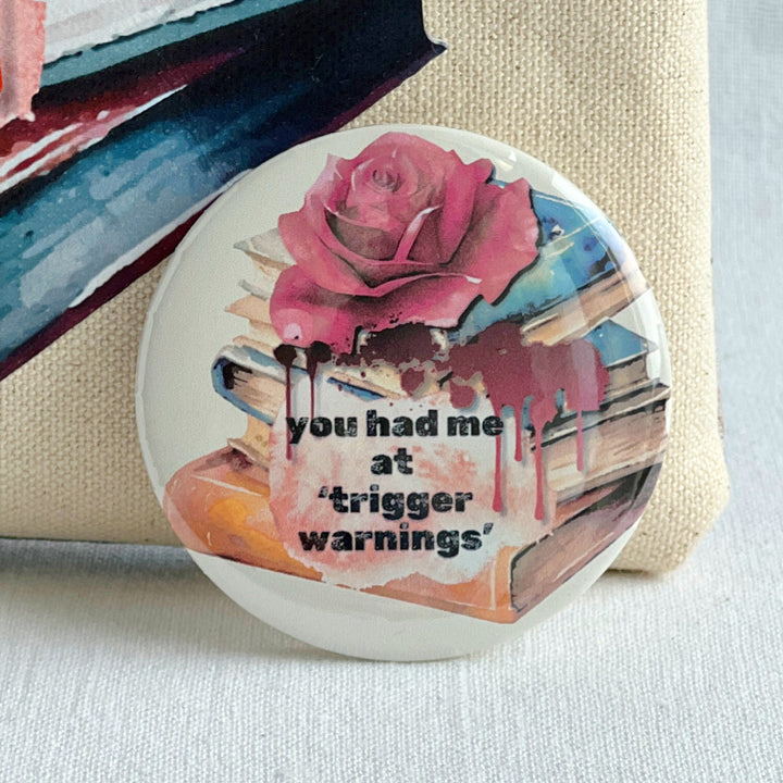 Round pinback button with a white background, a stack of colorful books with a rose on top and some blood dripping with the words, "You had me at trigger warnings."