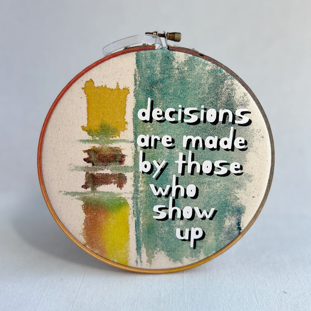 seven inch painted hoop with painted fabric in colors of yellow and mayan blues, with the words, decisions are made by those who show up.