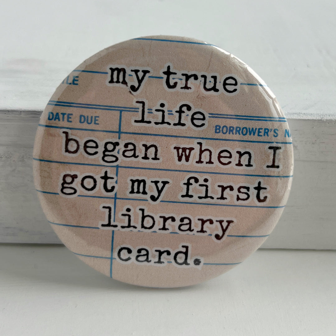 round pinback button with a library checkout card background and the words, "my true life began when I got my first library card."