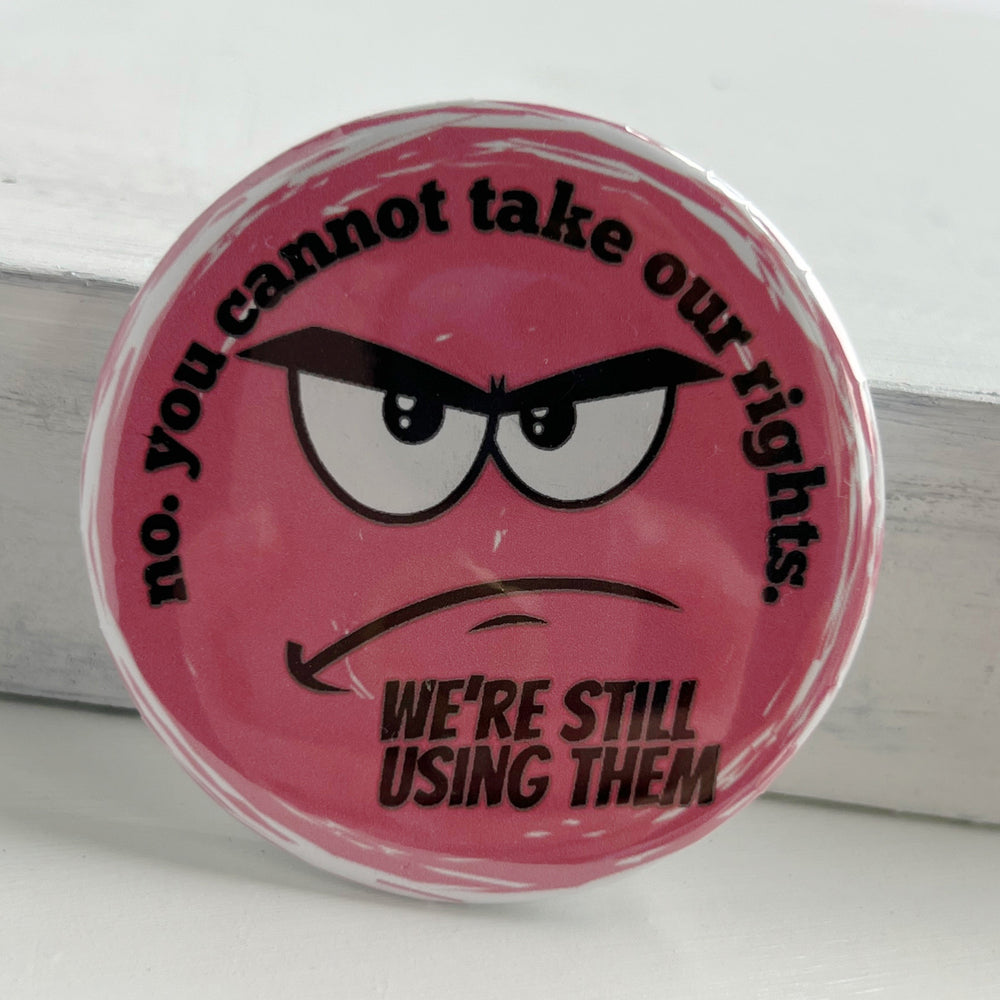 round pinback button with a pink background and the words, "no. you cannot take our rights. We're still using them."  The words surround an angry face.