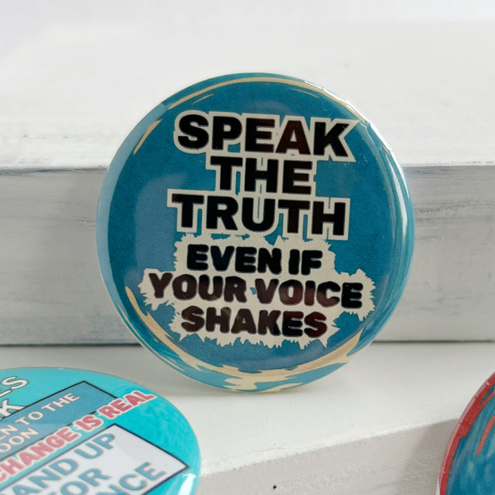 round, blue pinback button on a white background with the words, "speak the truth, even if your voice shakes."