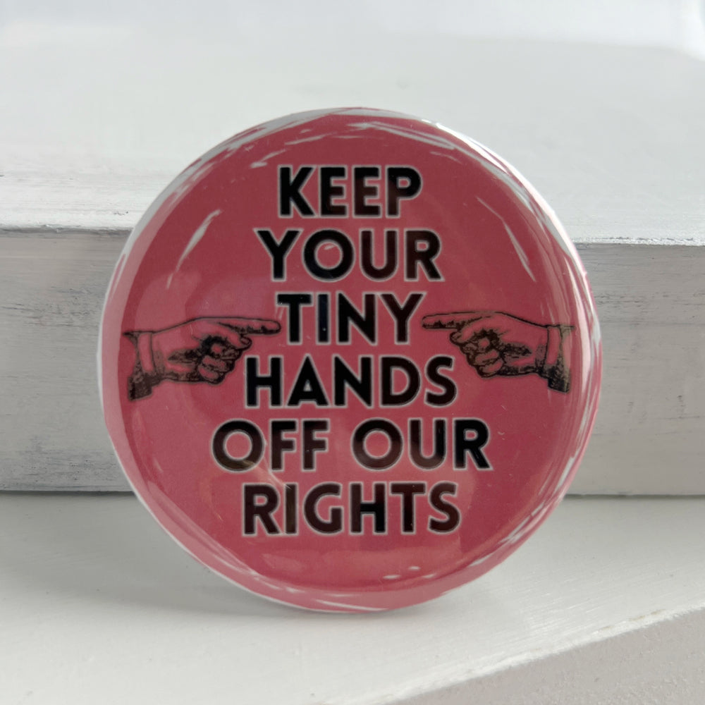 round pinback button with a pink background and the words, "Keep your tiny hands off our rights." a pair of hands in black point at the words.