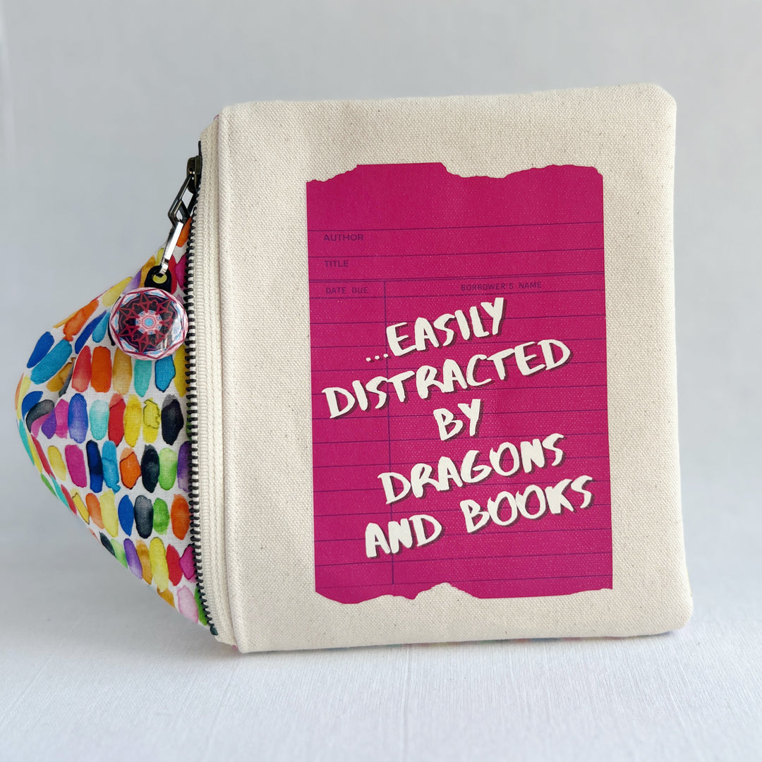 padded canvas zip bag with a coral red library card and the words, easily distracted by books and dragons, plus a colorful zipper pull and lining