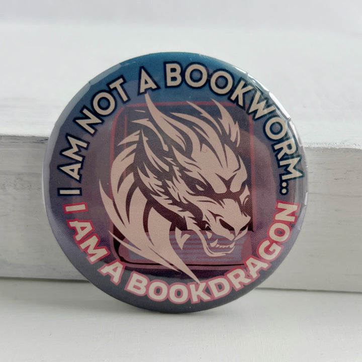 round pinback button with an ombre colored background with blues/pinks/purples, with the words, "I am not a bookworm..I am a book dragon" surrounding a light pink dragon figure on top of a book.