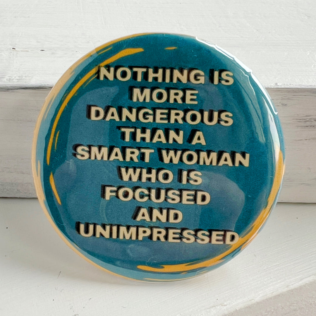 round pinback button with a blue backgroun, yellow streaks and the words, "Nothing is more dangerous than a smart woman who is focused and unimpressed."