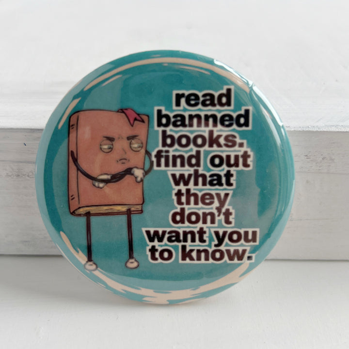 round pinback button with a teal blue background, a cartoon figure of a book with an angry face, and the words, "read banned book. find out what they don't want you to know."