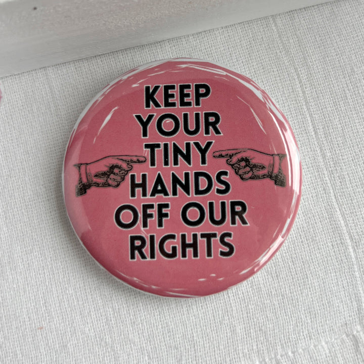 round pinback button with a pink background and the words, "Keep your tiny hands off our rights." a pair of hands in black point at the words.