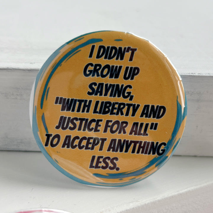 round pinback button with a yellow background with green streaks, and the words, "I didn't grow up saying, "with liberty and justice for all" to accept anything less."