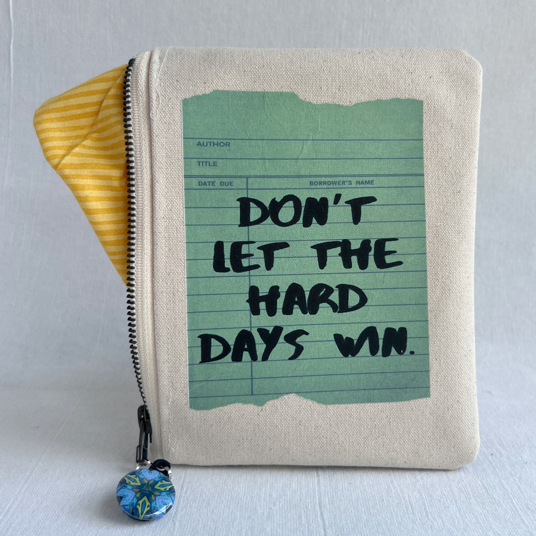 padded canvas zip bag with a green library card and the words, Don't Let the Hard Days Win, plus a colorful zipper pull and lining