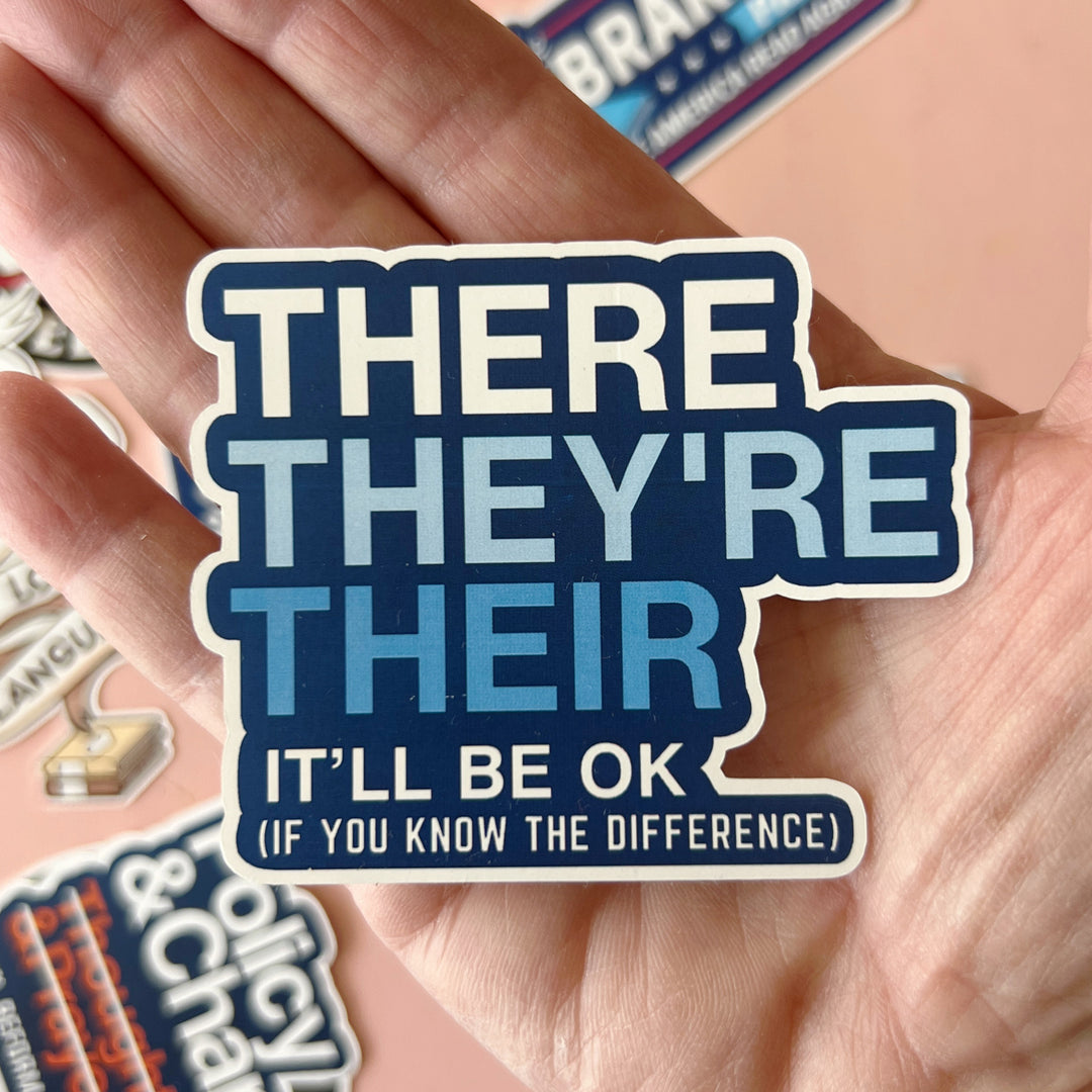 A word sticker with blue background and white lettering, with the words, "There They're Their. It'll be ok. If you know the difference.' A hand is holding the sticker.