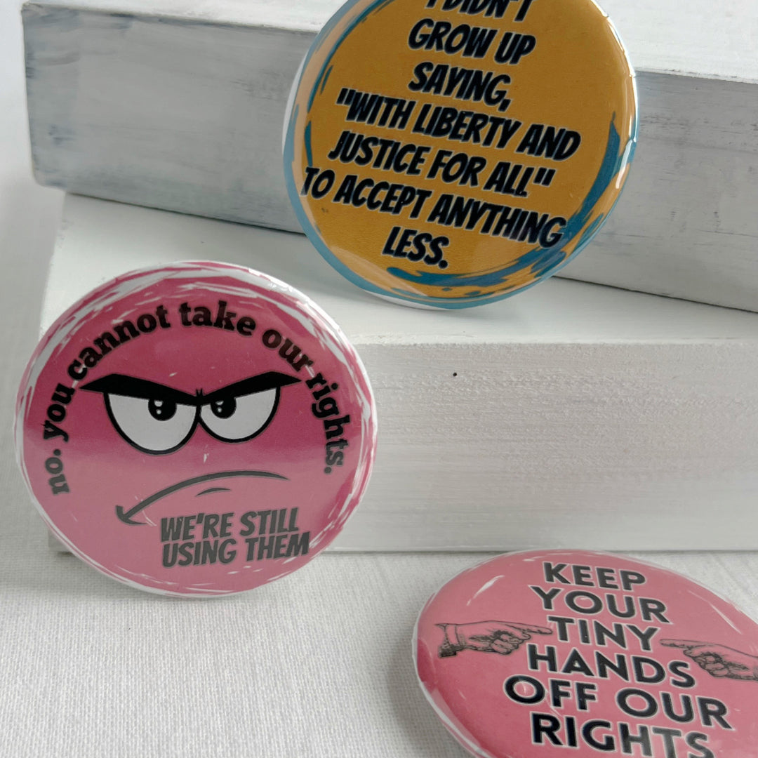 round pinback button with a pink background and the words, "no. you cannot take our rights. We're still using them."  The words surround an angry face. Two other buttons are also pictured.