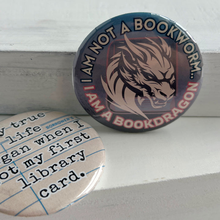 round pinback button with an ombre colored background with blues/pinks/purples, with the words, "I am not a bookworm..I am a book dragon" surrounding a light pink dragon figure on top of a book.