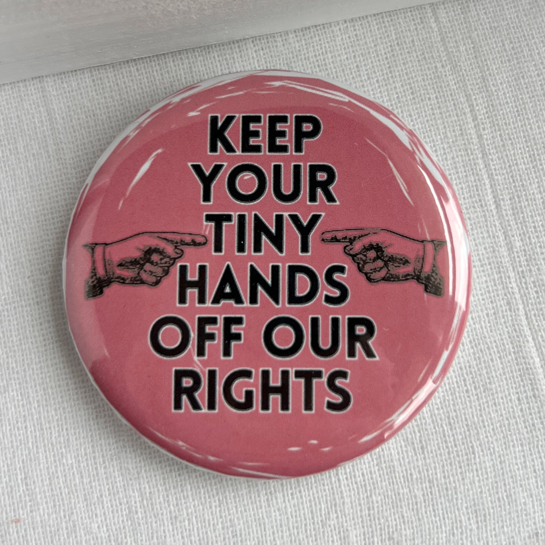 round pinback button with a pink background and the words, "Keep your tiny hands off our rights." a pair of hands in black point at the words.