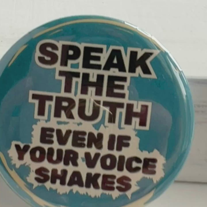 round, blue pinback button on a white background with the words, "speak the truth, even if your voice shakes."