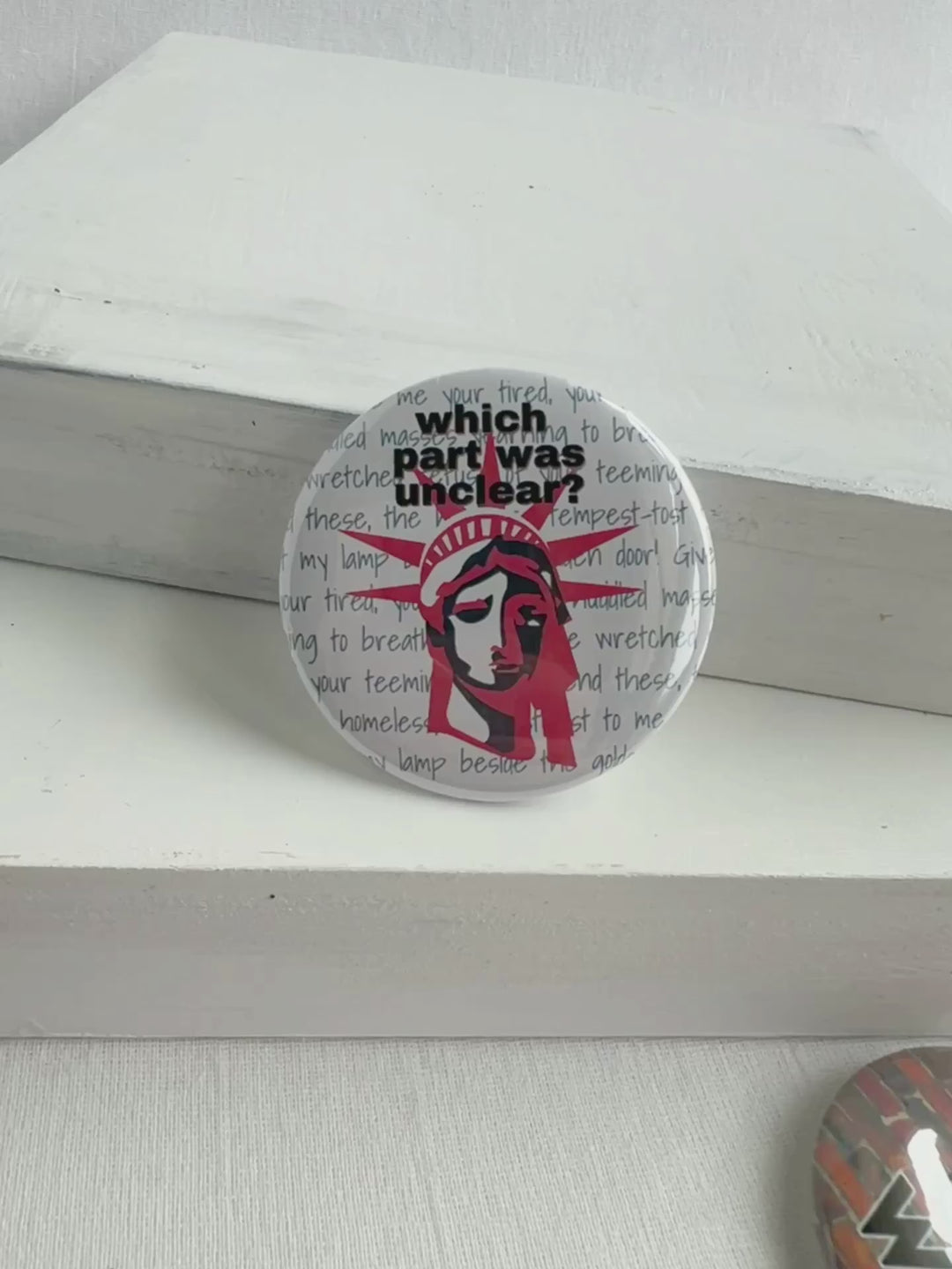 round pinback button with a white background, grey words spelling out some of the words on the base of Lady Liberty, such as give me your poor, your tired, your huddle masses. The words, "which part was unclear " label the top of the button, with a picture of Lady Liberty's head in red and blue.